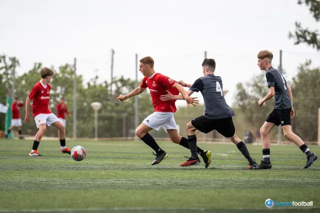 Youth soccer players on a green field: a player in red dribbles past a defender in black.