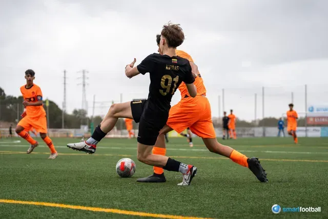 Soccer players in black and orange uniforms on a green field. One player attempts to kick the ball as another defends.