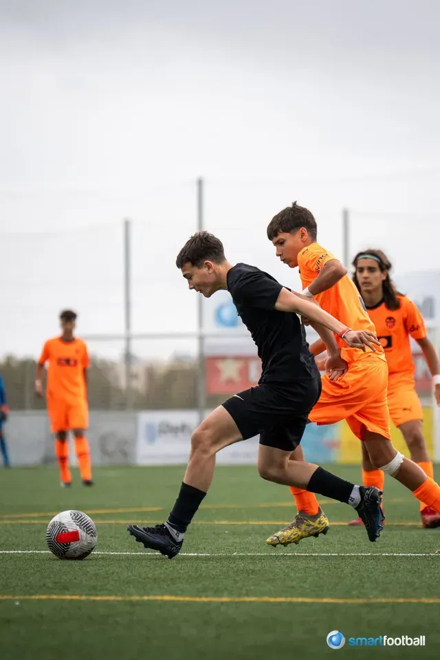 Soccer players in black and orange uniforms on a green field. A player in black dribbles past an orange-clad opponent.