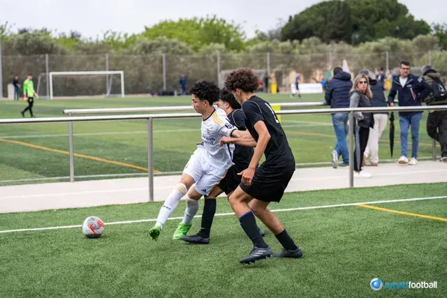 Soccer players one in white dribbles the ball, two others in black attempt to stop him. Spectators watch in the background.