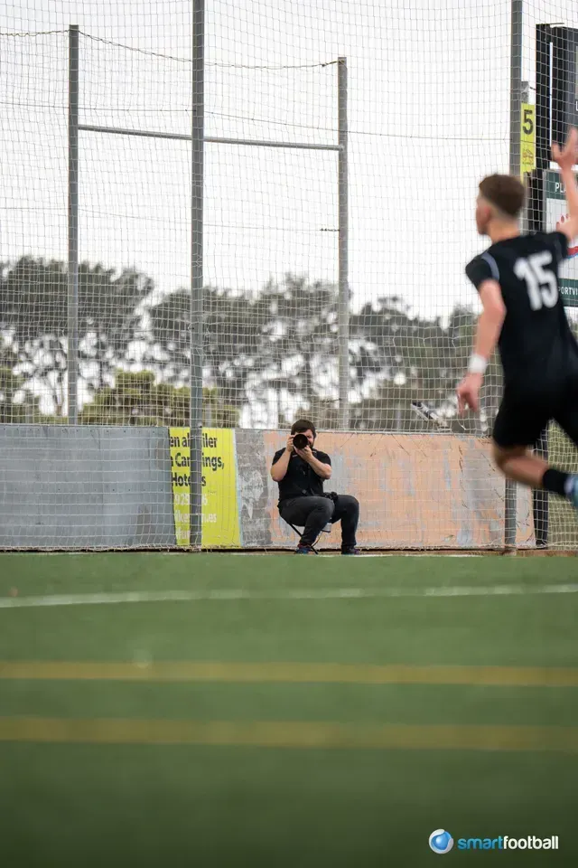 Photographer crouches at a soccer field, taking a picture of a player in black uniform.