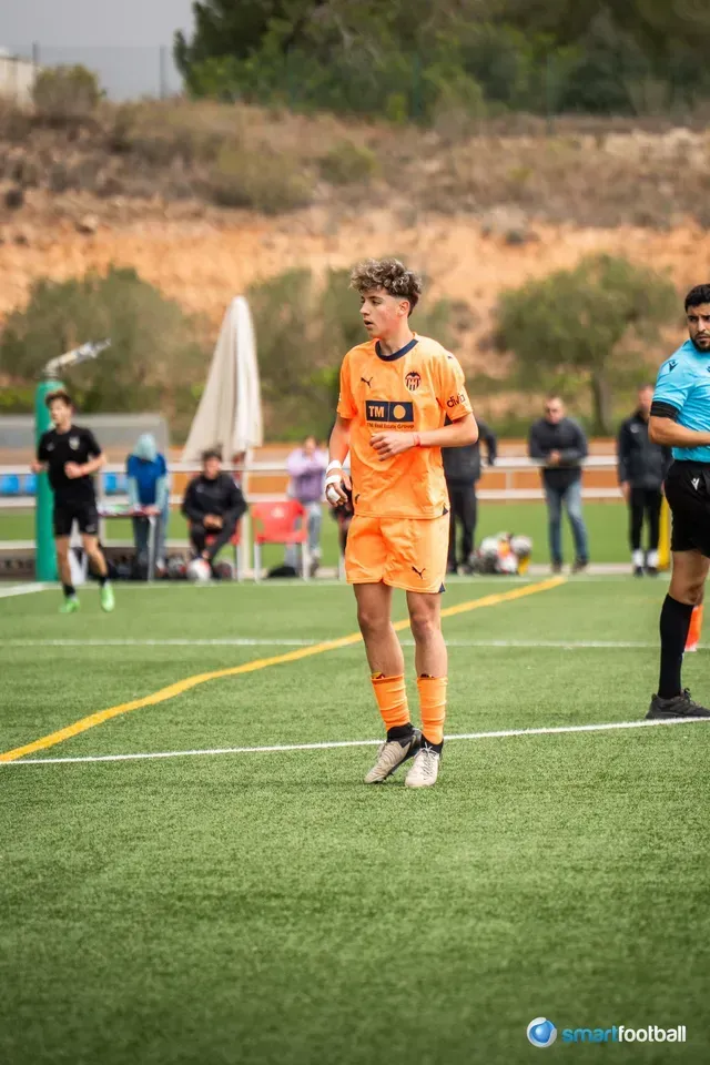 Soccer player in orange uniform stands on green field, eyes ahead, near the sideline.