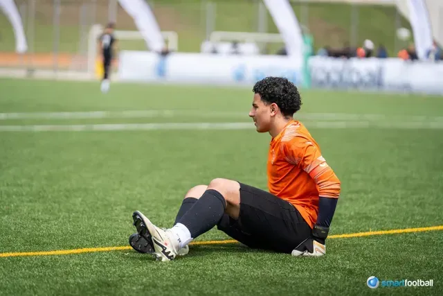 Soccer player in orange shirt and black shorts sits on field, observing the game.