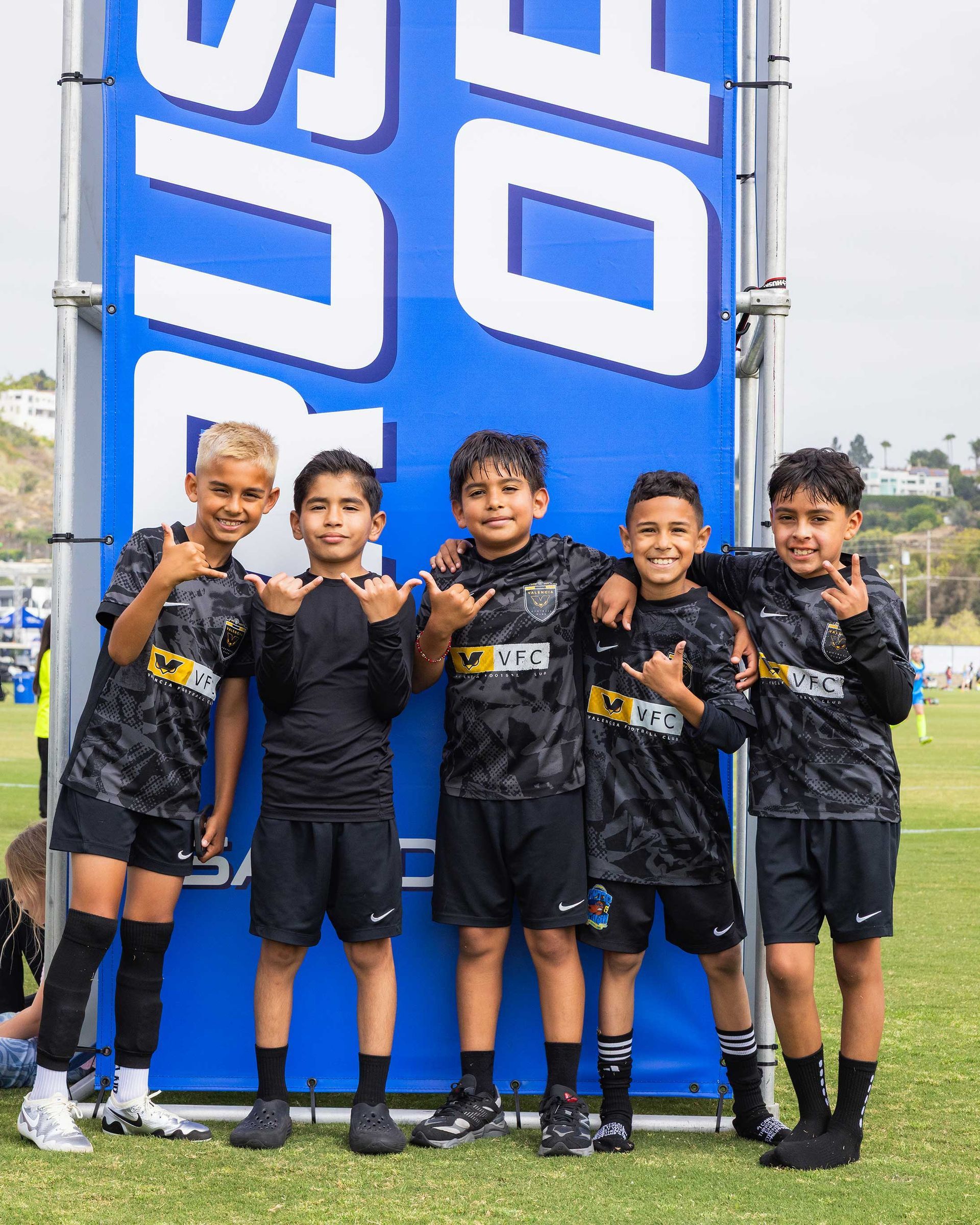 Five kids in black sports uniforms pose smiling beside a large blue banner on a grassy field.