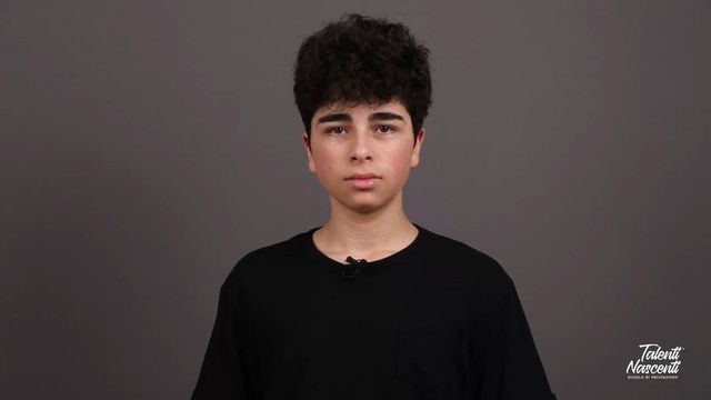 Adolescente con capelli scuri e ricci, maglietta nera su sfondo grigio, che guarda la telecamera.