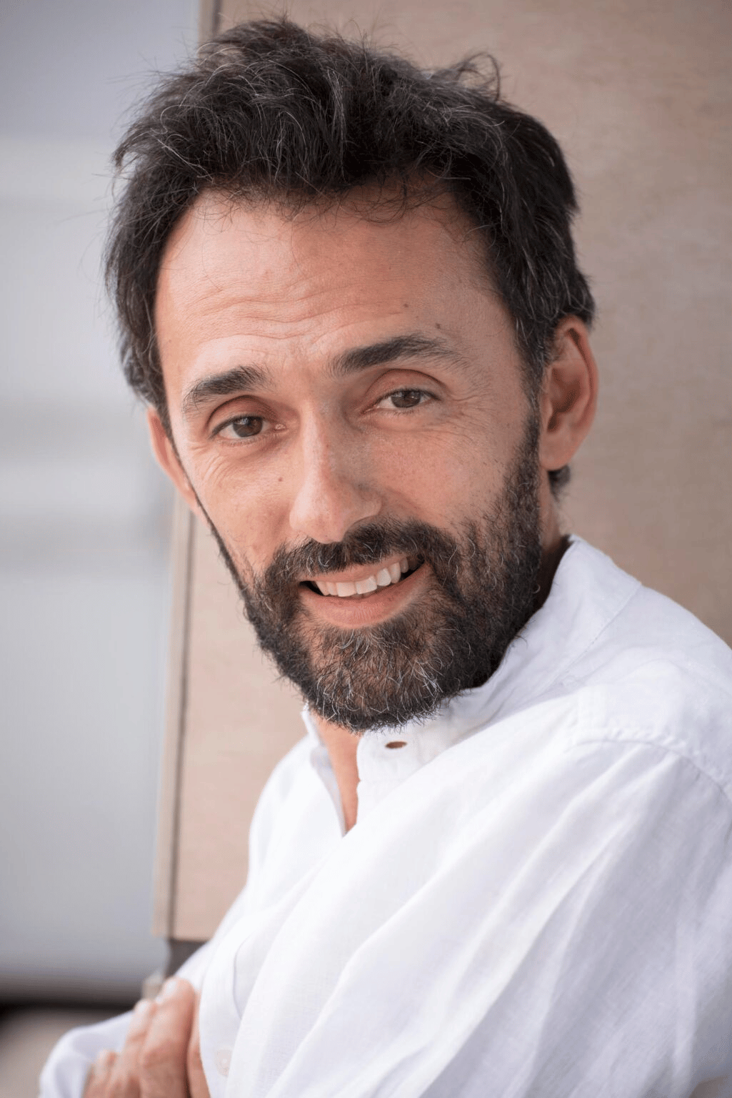 Un uomo con la barba sorride alla telecamera, indossa una camicia bianca ed è appoggiato a un edificio.