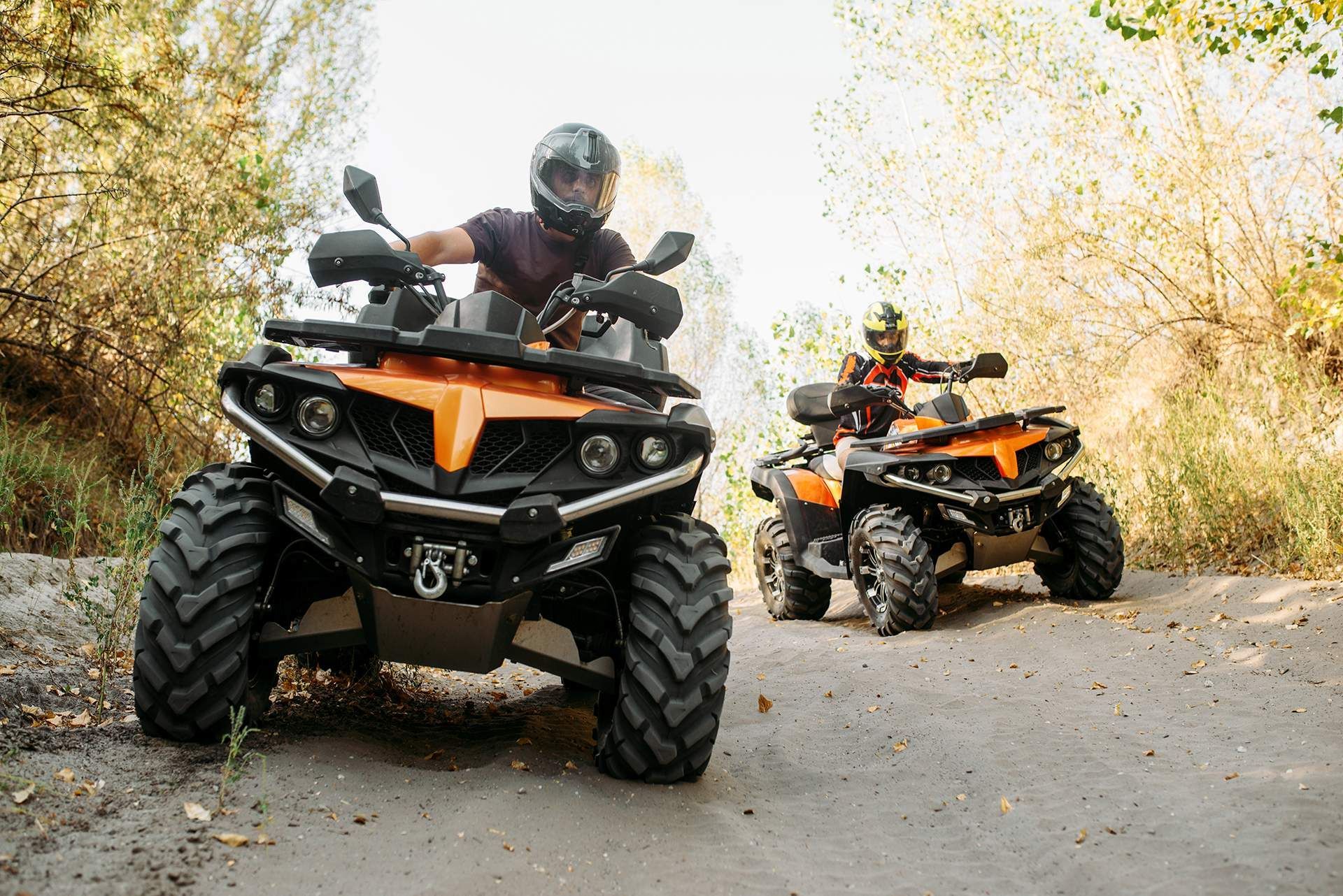 Two riders on orange ATVs on a wooded dirt path, one close in front and one behind.