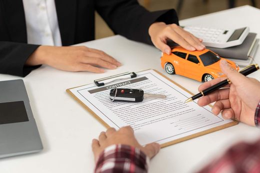 Hands reviewing a car damage report with a toy orange car and clipboard at a desk