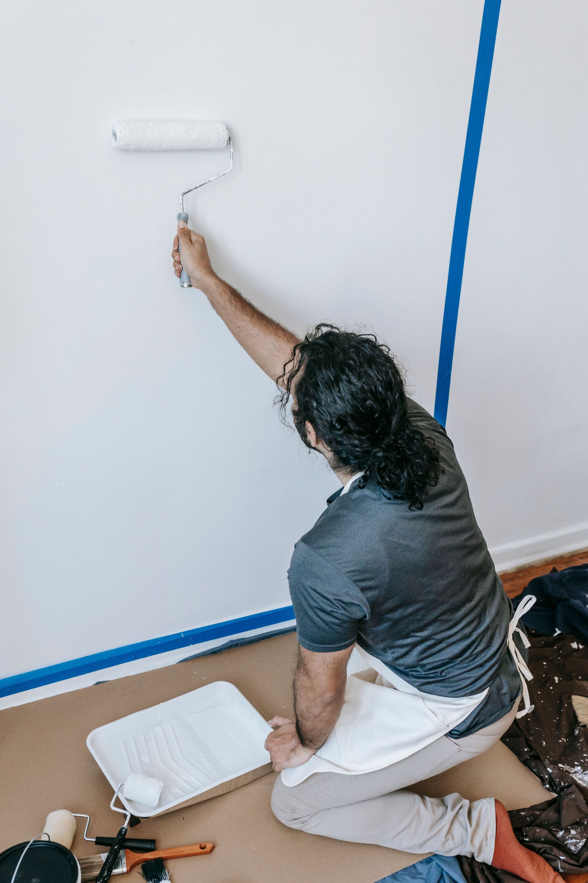 Person kneeling and painting a white wall with a roller, blue tape marking the edges.