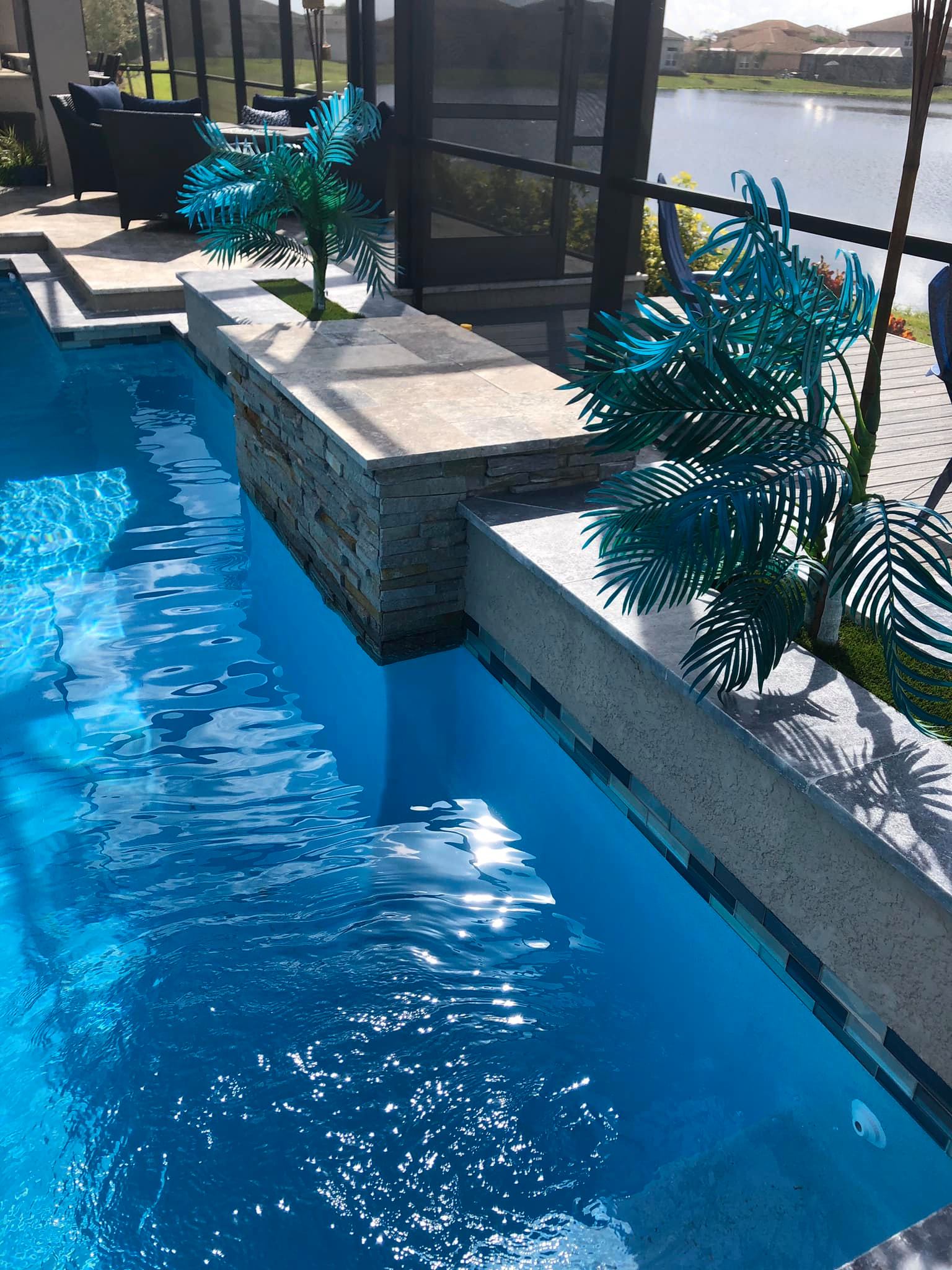 Pool with blue water, a stone wall, and turquoise palm tree sculptures, near a lake.
