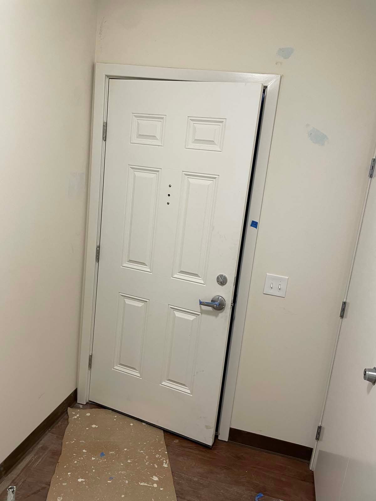 Partially open white front door in an unfinished room with beige walls and a bare floor.