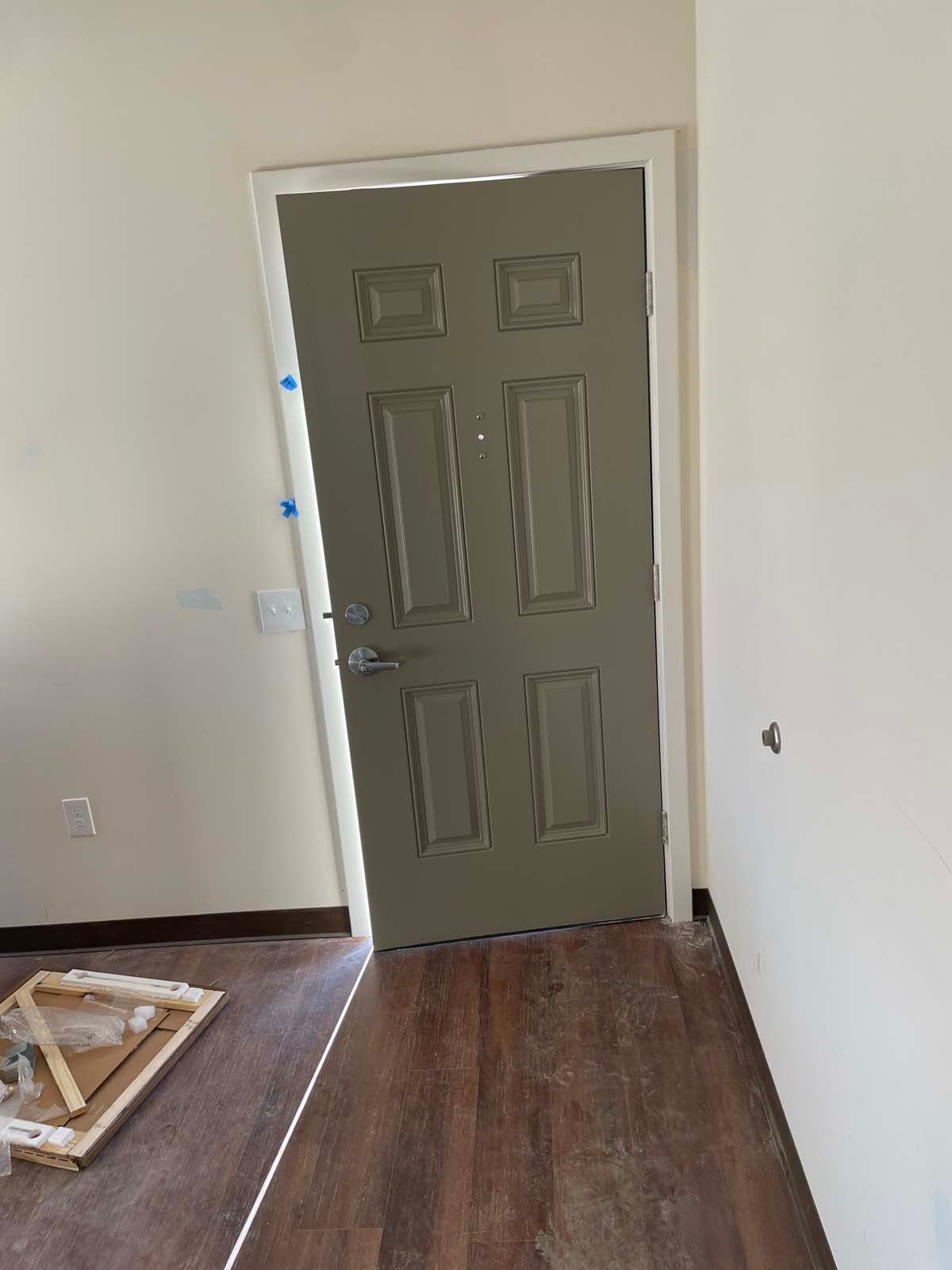 Empty room with a closed gray front door and wood flooring, with a fallen frame in the corner.