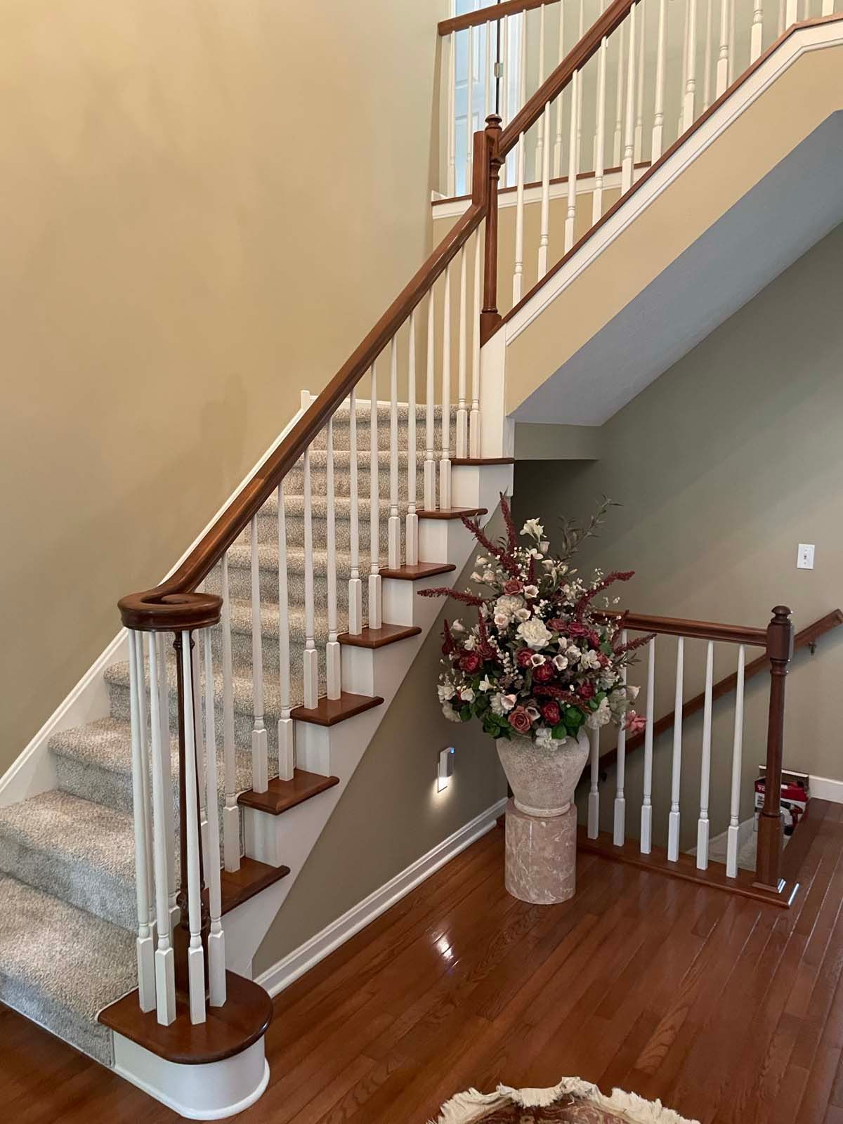 Staircase with white balusters and wood handrails beside a vase of flowers on a hardwood floor.