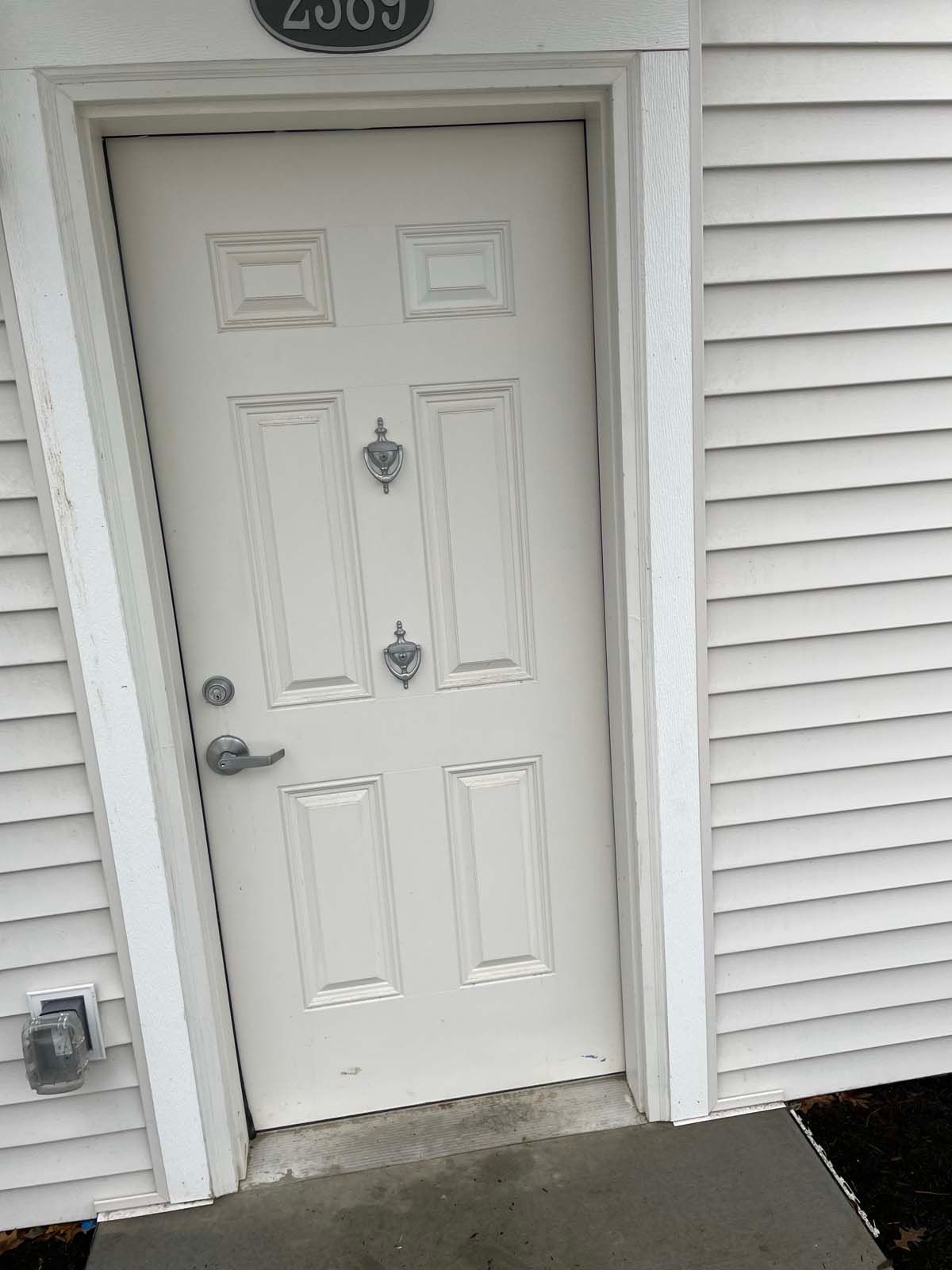 White front door with decorative panels and a small wreath on a beige-sided house entryway.