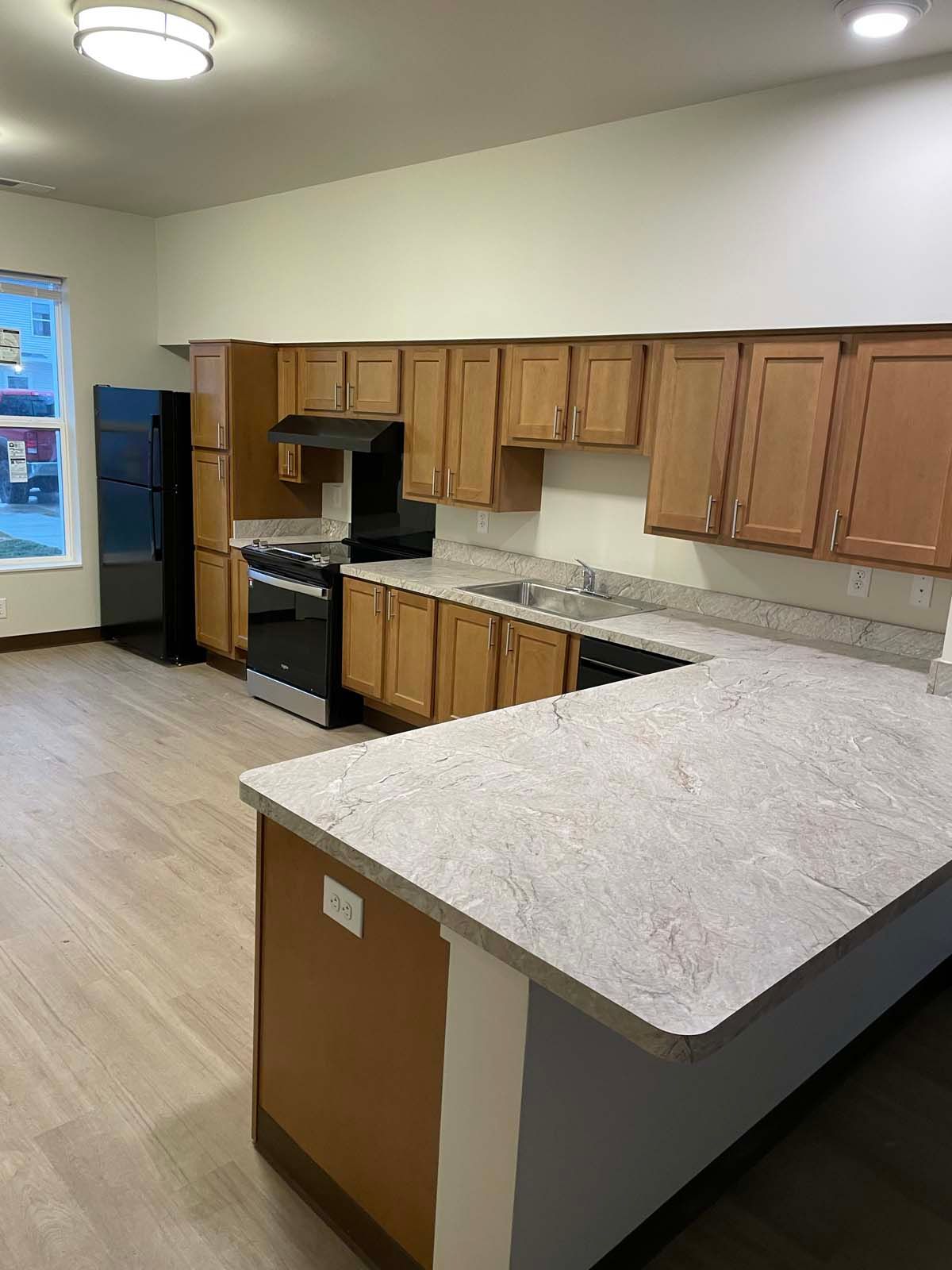 Empty kitchen with wooden cabinets, speckled countertops, black stove, and light wood flooring.