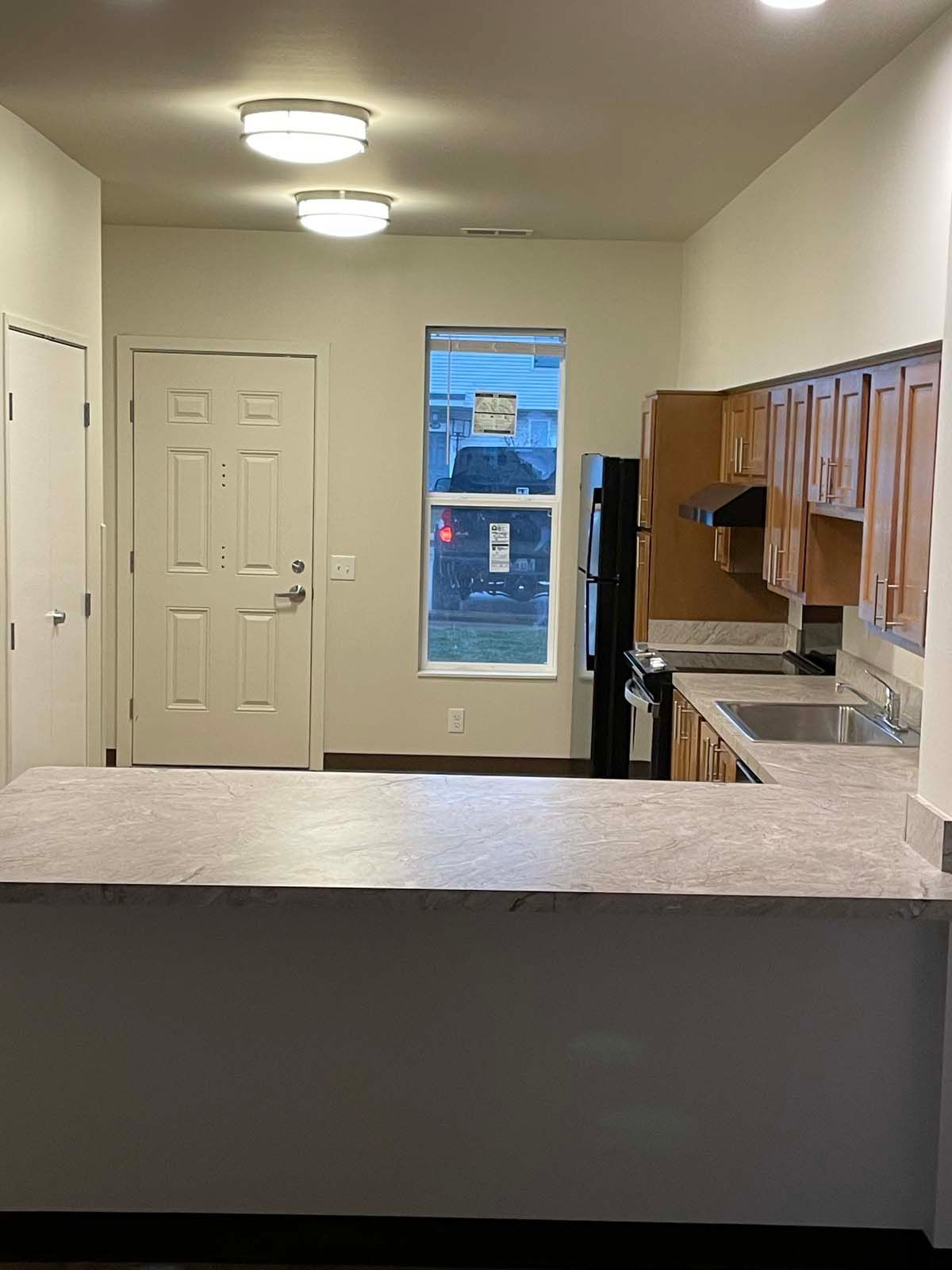 Small kitchen with white cabinets, counter, ceiling lights, and a window at the far end.