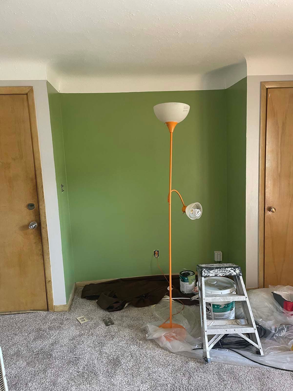 Room with green accent wall, two wooden doors, floor lamp, step stool, and renovation supplies on carpet.