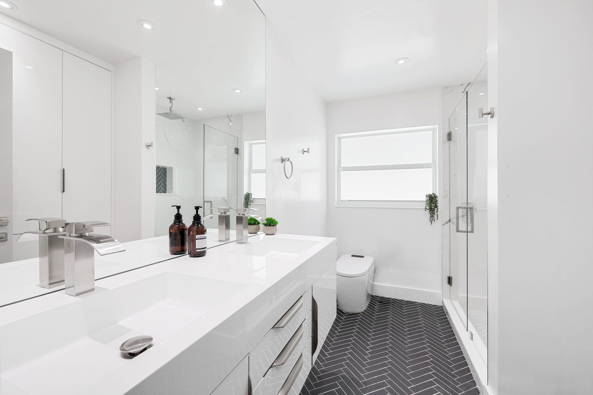 Bright white modern bathroom with double vanity, large mirror, toilet, and patterned dark floor tile.