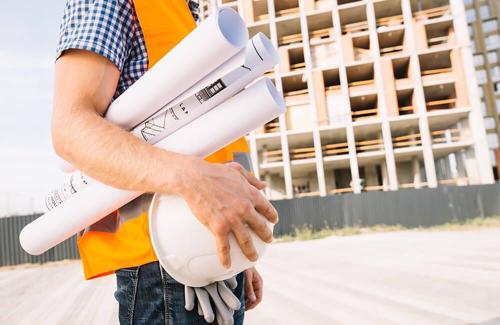 A construction worker is holding a hard hat and blueprints at a construction site.