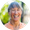 Smiling person with short gray-blue hair in a peach shirt against a softly blurred green outdoor background