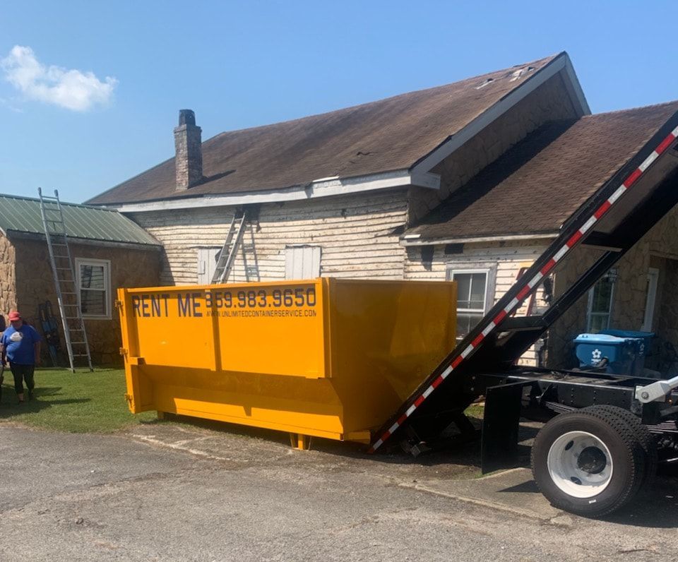A yellow dumpster on a truck bed is parked in front of a weathered building.