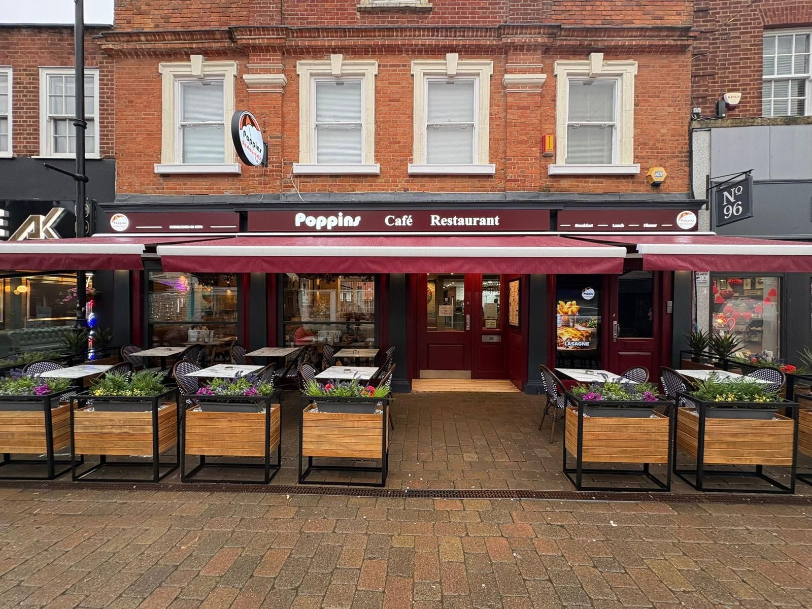Front view of a red-brick café with burgundy awning and outdoor seating on a cobblestone street
