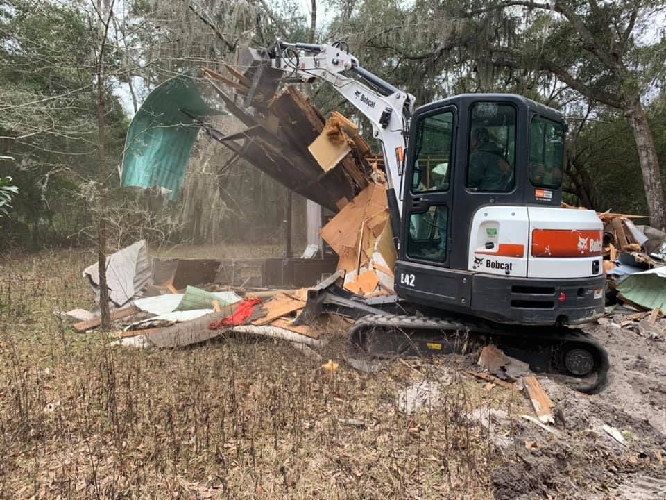 A small excavator is destroying a house in a field.