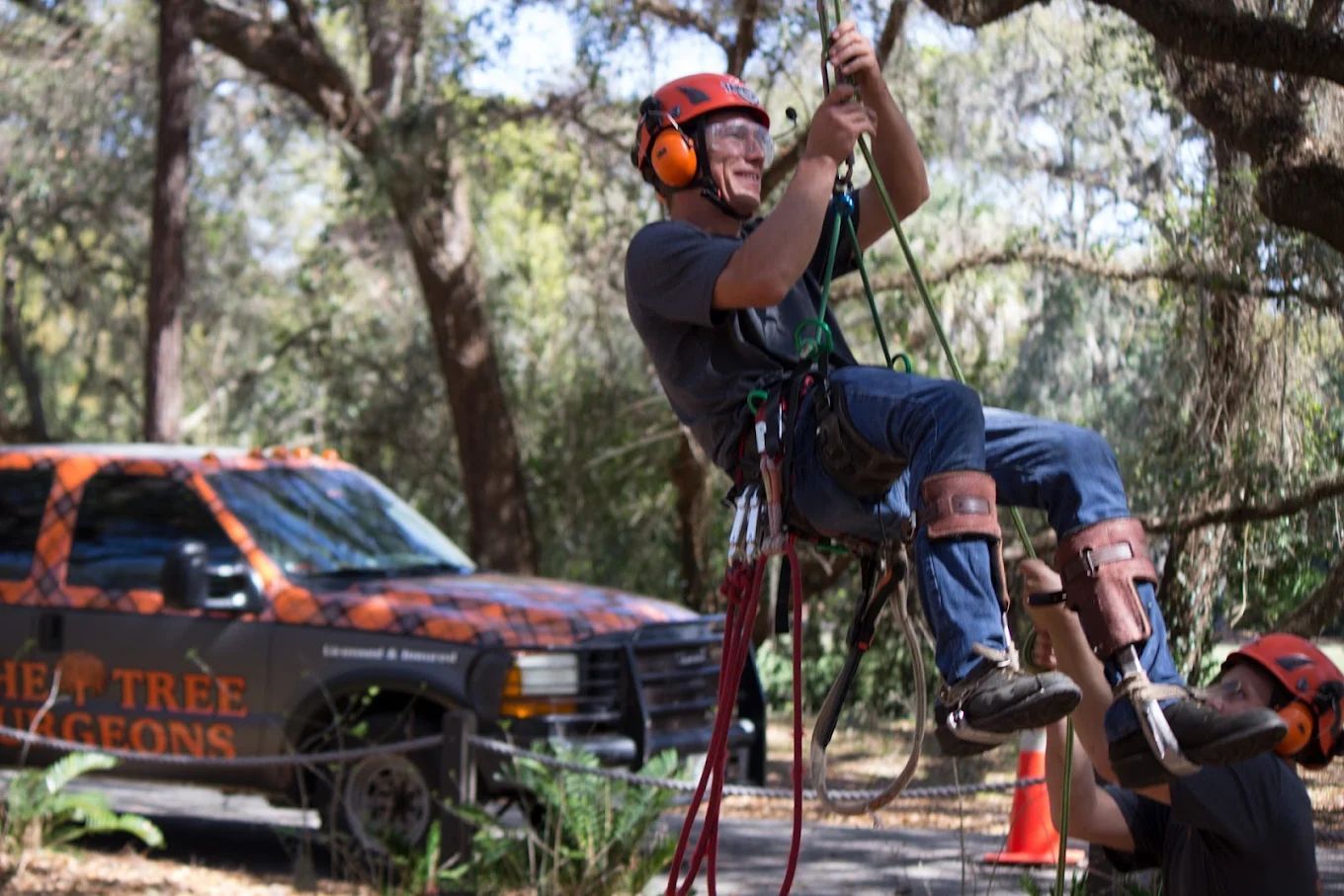 Rappelling climber in orange helmet descends a rope beside a parked vehicle in a wooded area