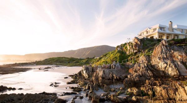 Coastal hotel on rocky cliff overlooking a beach, with mountains in the background during golden hour.