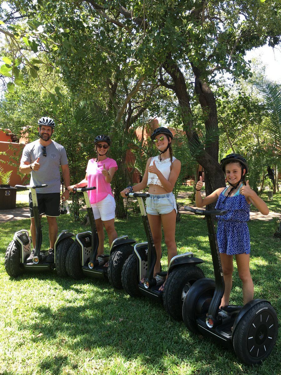 Family on Segways in a park, smiling and giving thumbs up. Sunlight filters through trees.