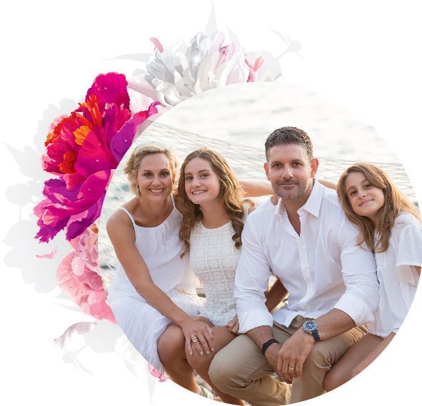 Family of four smiling together, dressed in white, posing outdoors. Bright pink flowers in corner.