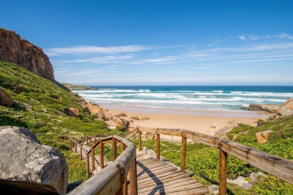 Wooden walkway leading down to a sandy beach and ocean under a blue sky.