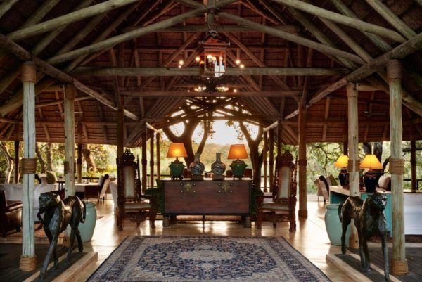 Lodge interior with wood beams, tables, antique furniture, and a rug, overlooking an outdoor setting.