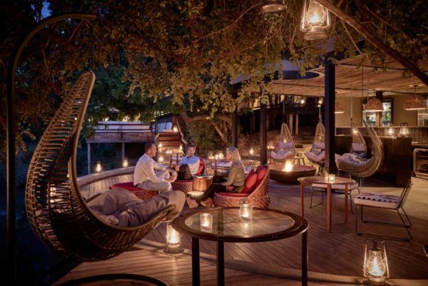 People relaxing outdoors at night, seated in wicker chairs, illuminated by lanterns and soft lighting.