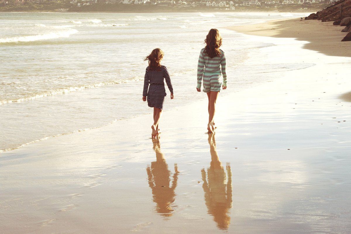 Two women walking on a sandy beach, leaving reflections in the wet sand; ocean waves and coastline in the distance.