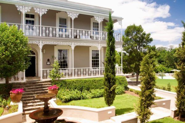 Elegant two-story house with balcony, fountain, and landscaped gardens, sunny day.