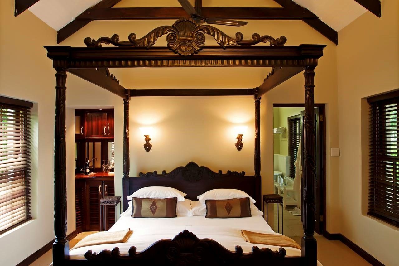 Bedroom with dark wooden four-poster bed, two sconces, and a small bar in a light-colored room.