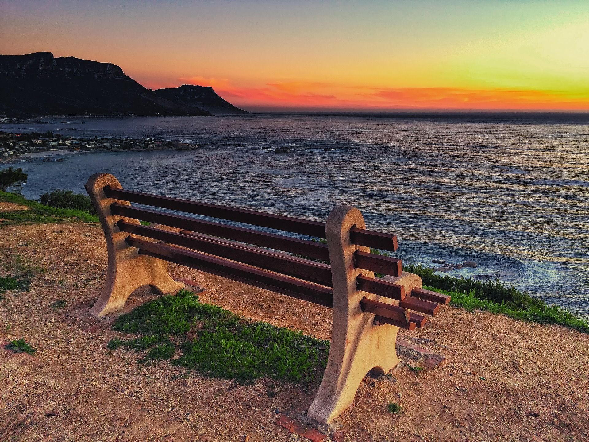 Bench overlooking the ocean at sunset. Orange and yellow sky, dark mountain in background.