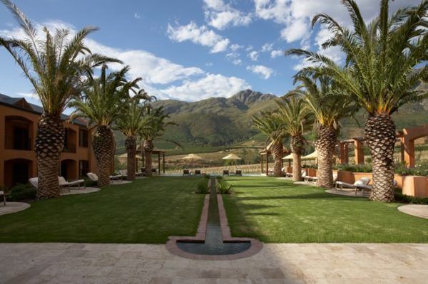 Lush green lawn flanked by palm trees and a low building, with mountains in the distance under a blue sky.