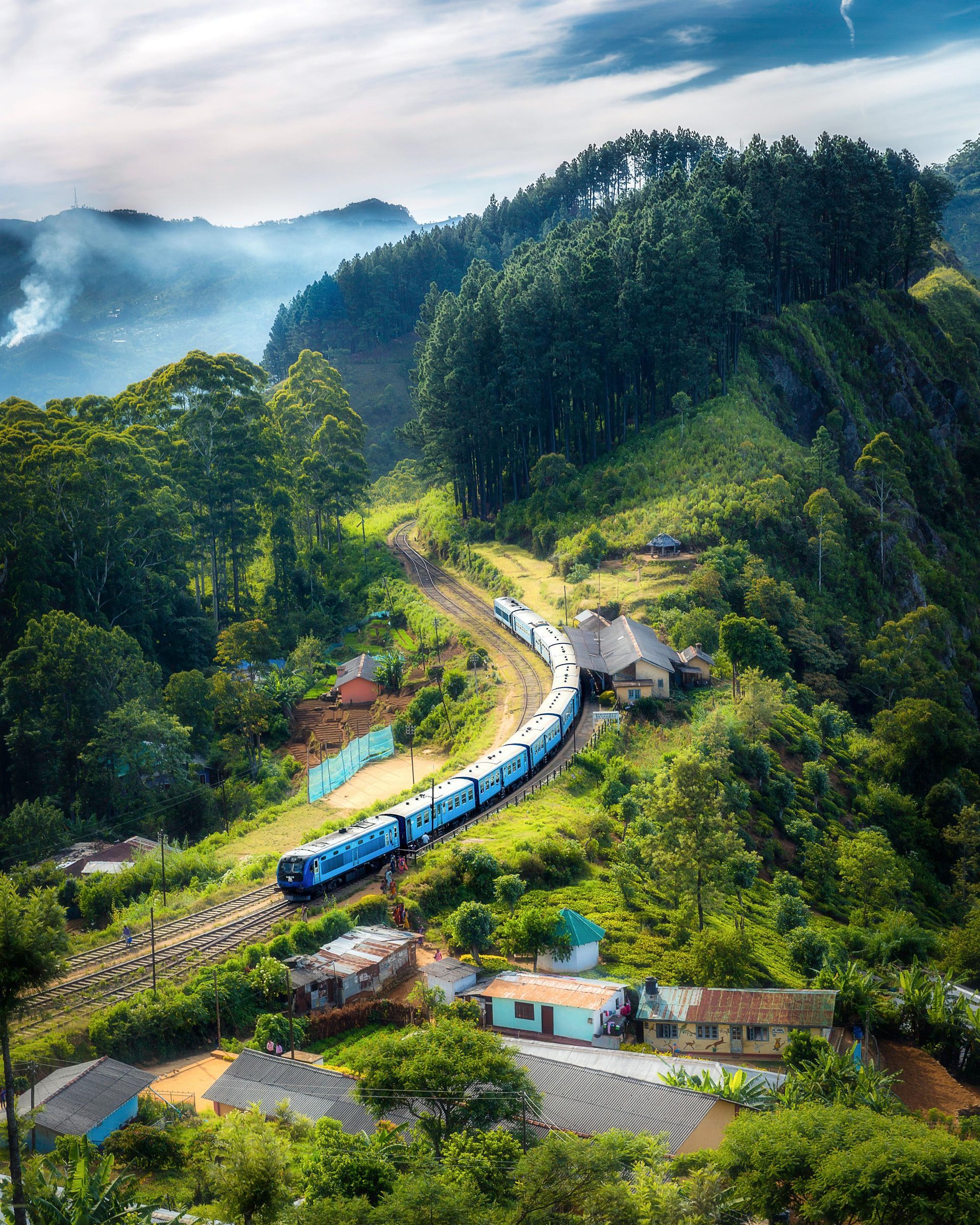 Train winding through lush green hills, passing small houses and a tunnel entrance in the mountains.