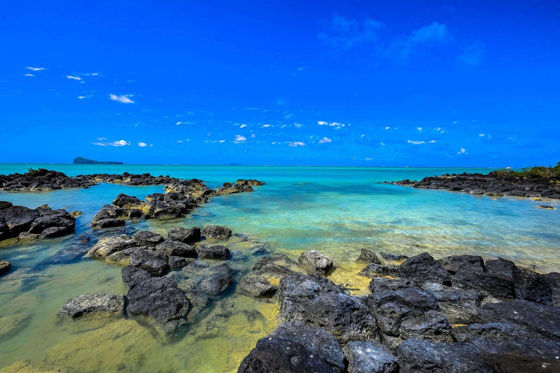 Clear turquoise water over black rocks under a bright blue sky; a tropical coastal scene.