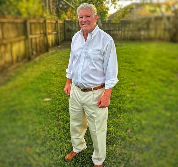Man in a white shirt and beige pants standing on a grassy lawn by a wooden fence