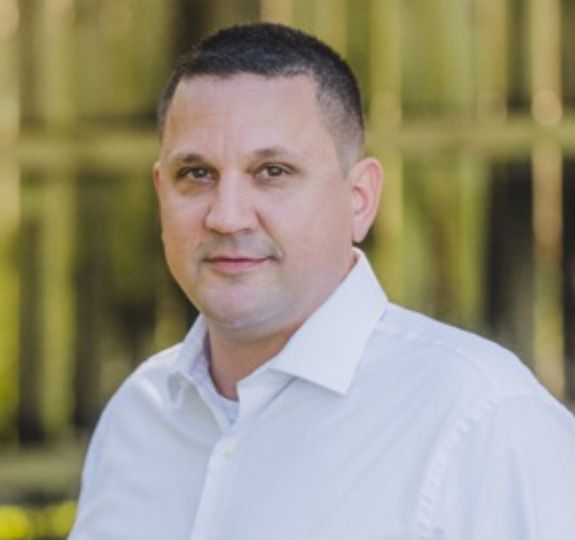 Portrait of a man in a white shirt standing outdoors against a blurred fence background