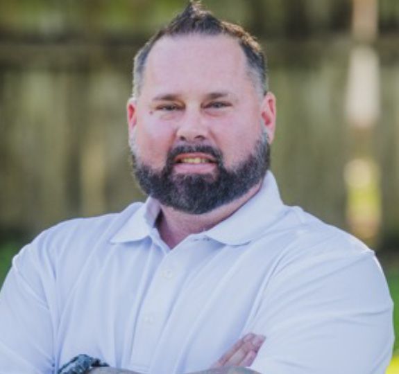 Smiling bearded man in a white polo shirt standing outdoors with arms crossed