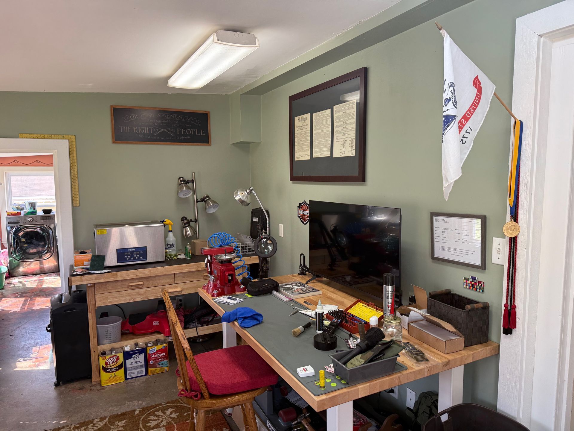 Cluttered home office with a desk, computer monitors, tools, and storage bins in a small room.