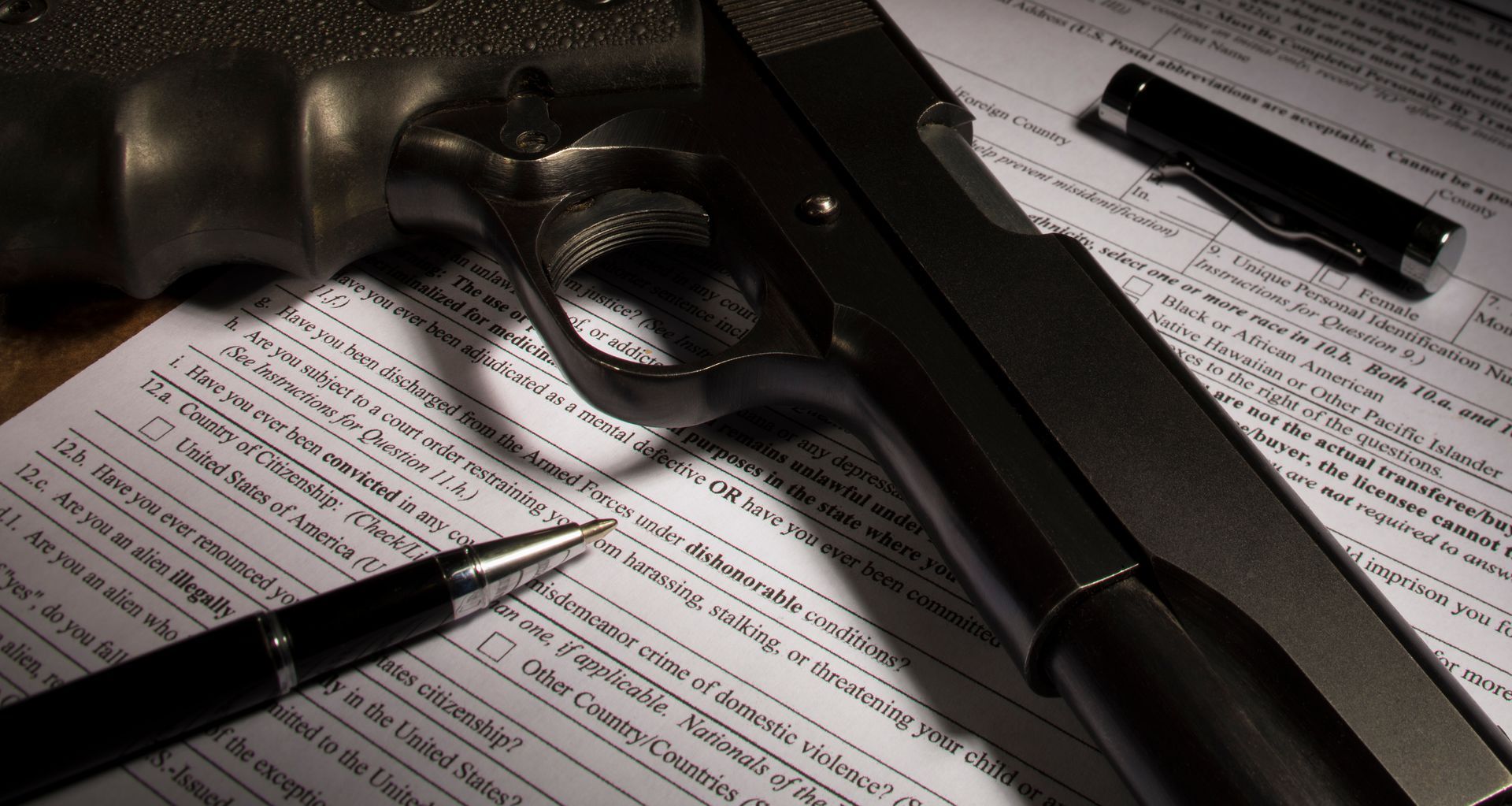 Black handgun, bullets, and pens on paperwork in a dimly lit desk scene