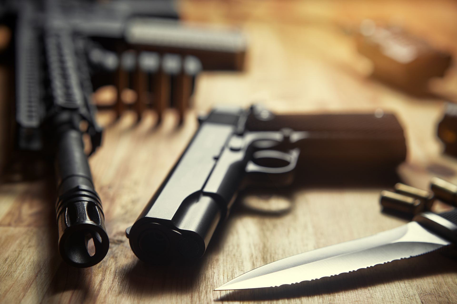 Handgun, knife, and ammunition on a wooden table, with a rifle in the background