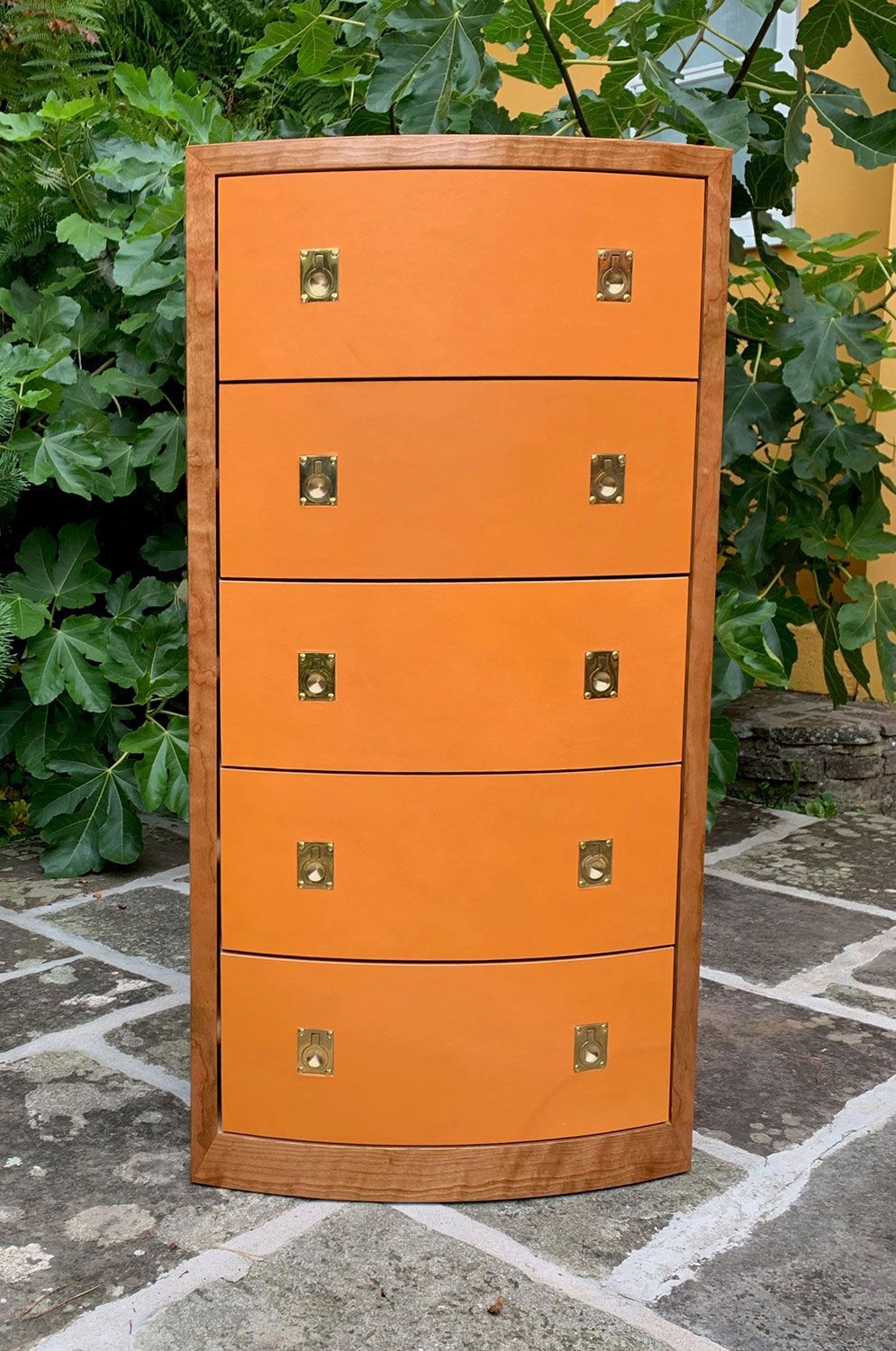 A hand crafted wooden chest of drawers with orange drawers and gold handles is sitting on a tiled floor.