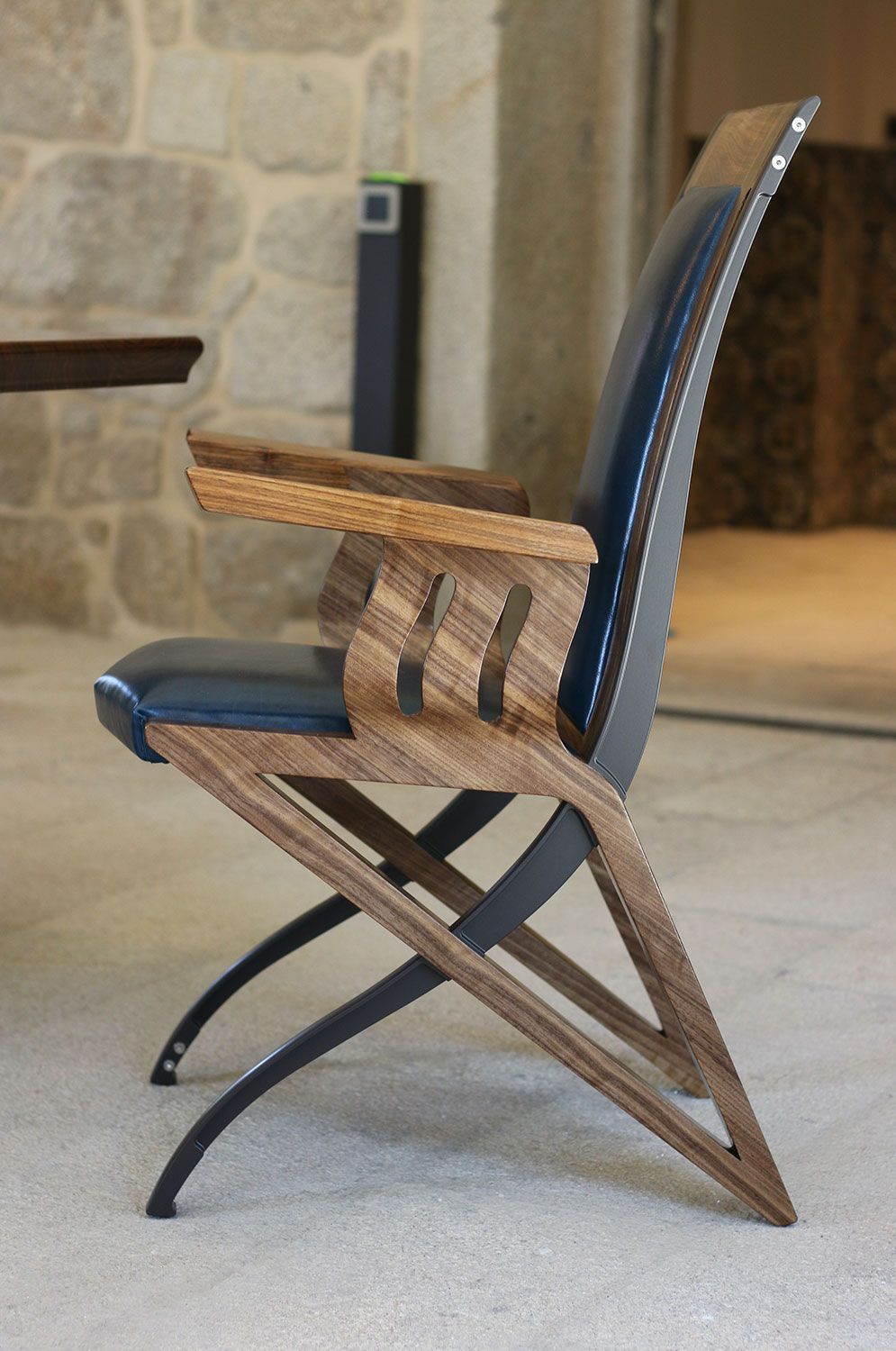 A wooden chair designed by sebastian blakeley with a blue leather seat sits in front of a stone wall
