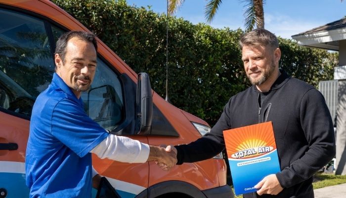 Two men shaking hands in front of an orange van, one holding a brochure with a sun logo.